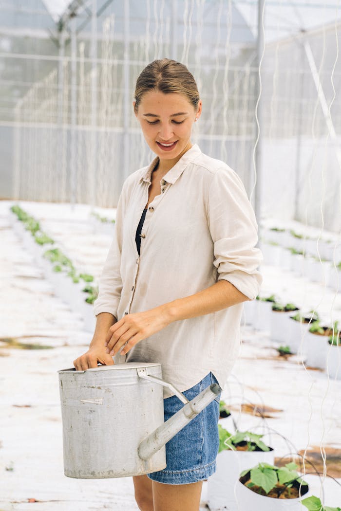 Smiling woman in a greenhouse with a watering can, nurturing plants for sustainable farming.
